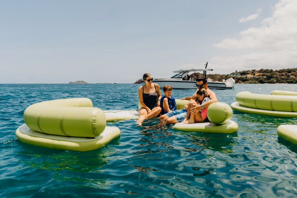 Family relaxing on floating platforms in turquoise water with Her Deal anchored nearby